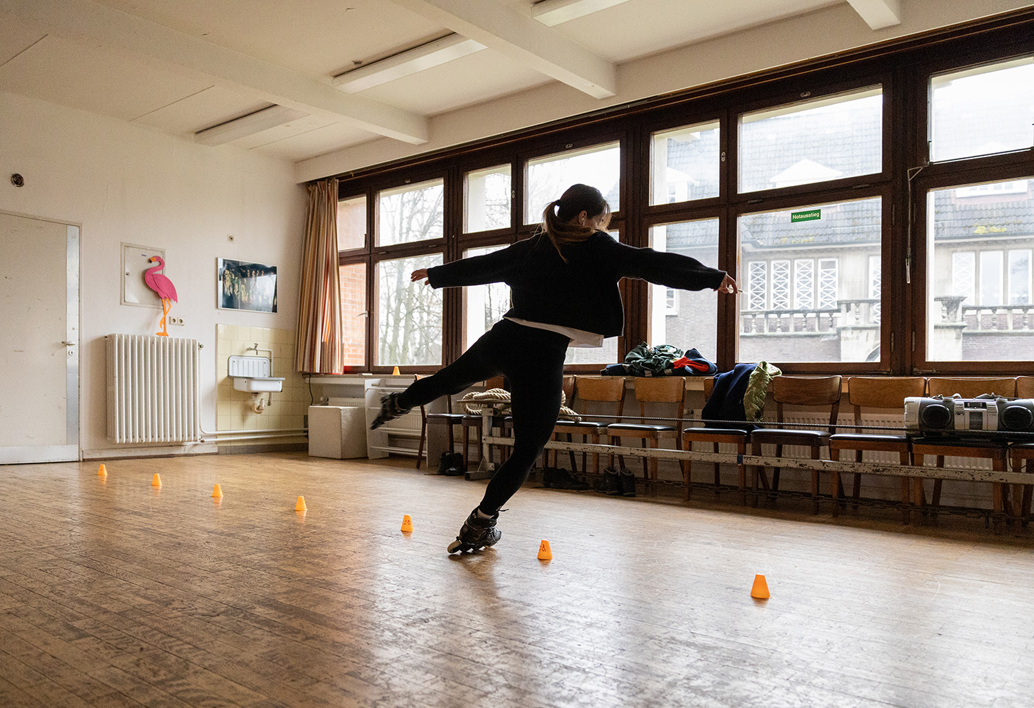 Junge Frau auf Inlineskater trainiert in einem großen Raum einer Schule. Auf dem Boden sind kleine Pylone aufgestellt durch die sie fährt.